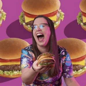 A girl holding a burger and smiling infront of a backdrop of burgers as an entry to be the Halifax Burger Bash Burger Ambassador